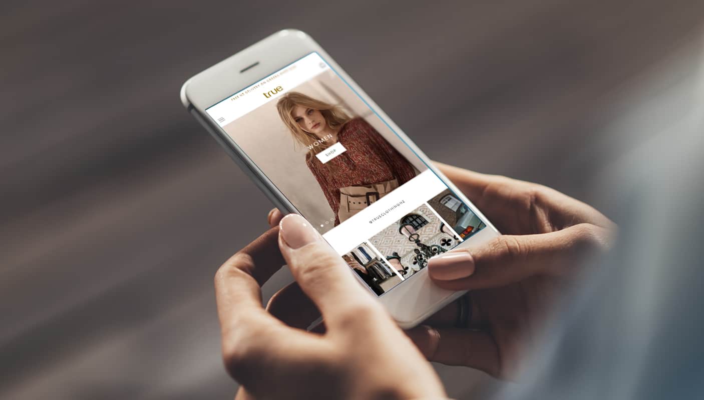 Woman with pale pink nail polish holding a white iPhone with a clothing ecommerce web design displayed